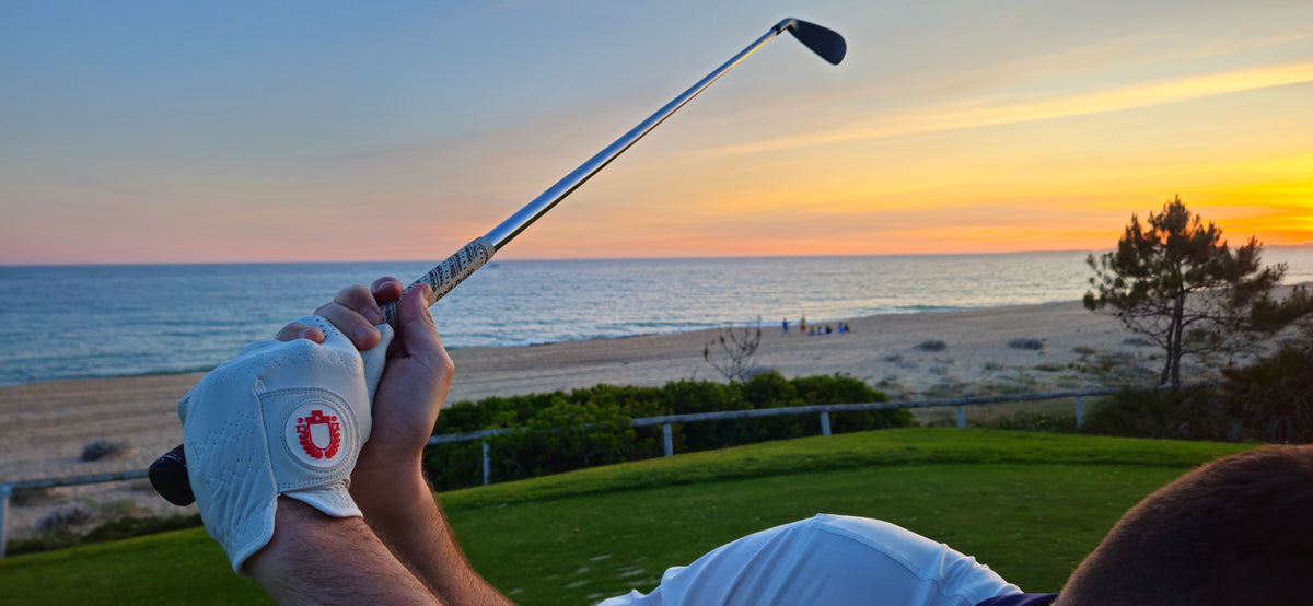 A close-up lifestyle shot of a golfer in a white golf glove holding a club, with a scenic ocean beach sunset background.